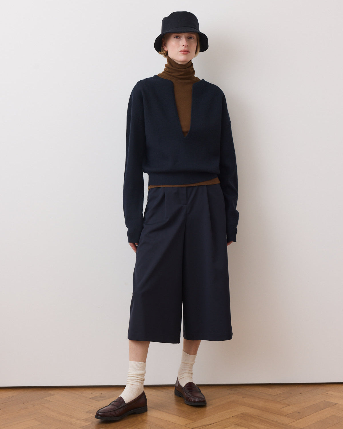 Person wearing a navy sweater, brown turtleneck, and black wide-leg pants against a white wall.