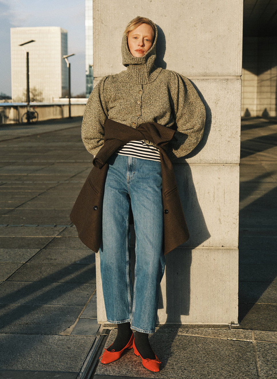 A woman stands outdoors against a concrete column wearing a textured olive green hooded sweater, a brown coat tied at the waist, blue jeans, black socks, and bright red shoes.