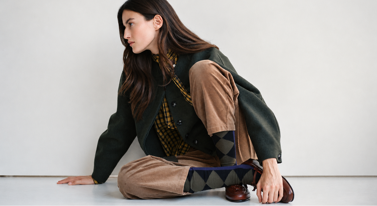 A model sits on the floor against a plain white wall, wearing a dark green jacket, brown trousers, a yellow and black argyle shirt, and brown leather shoes.